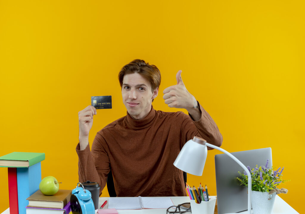 Young student holding credit card at desk with school tools, 